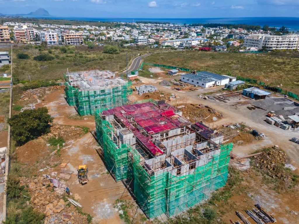 Drone photograph of the two Anna City blocks at full height under green scaffold netting, with Flic en Flac and the lagoon behind