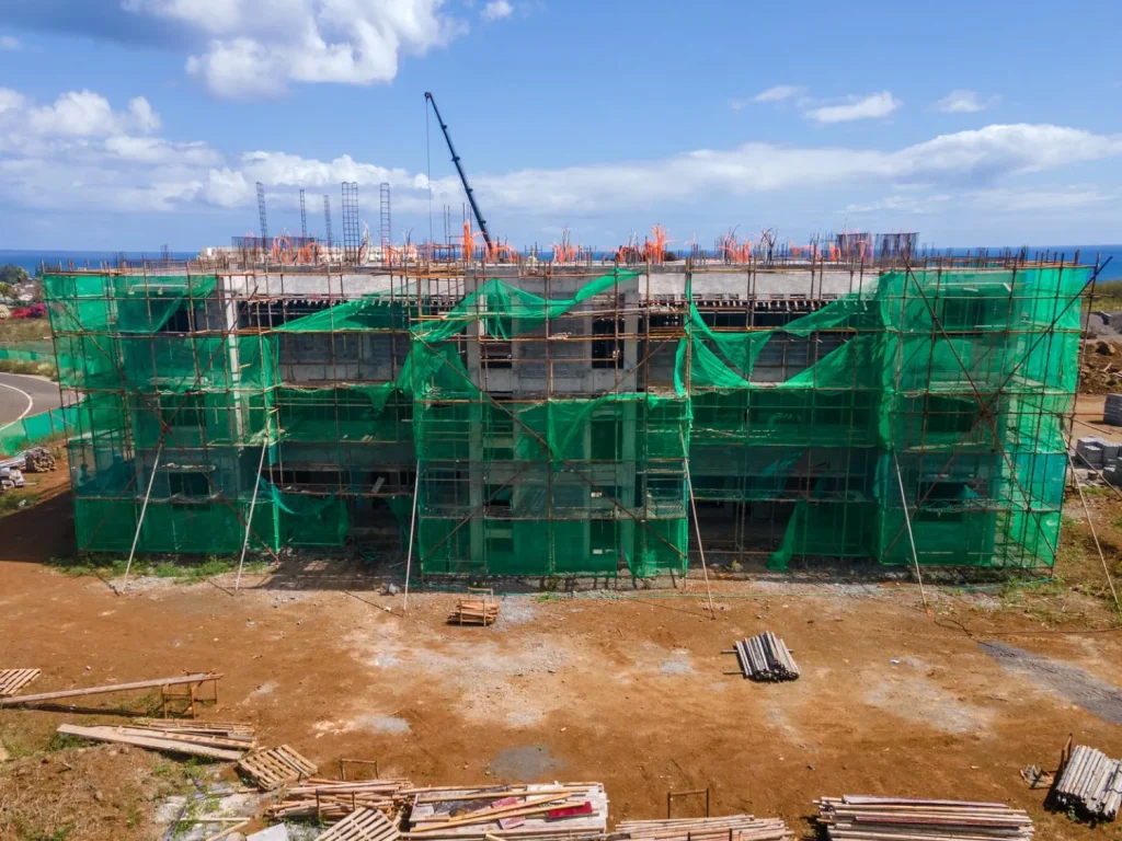 Frontal view of an Anna City block wrapped in green scaffold netting with a crane above and the sea behind